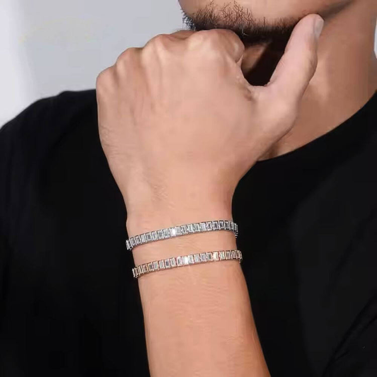 A person in a black shirt rests their chin on their hand, showcasing two sparkling Emerald Cut Moissanite Tennis Bracelets on their wrist. The focus is on the bracelets with only the lower part of the face partially visible.