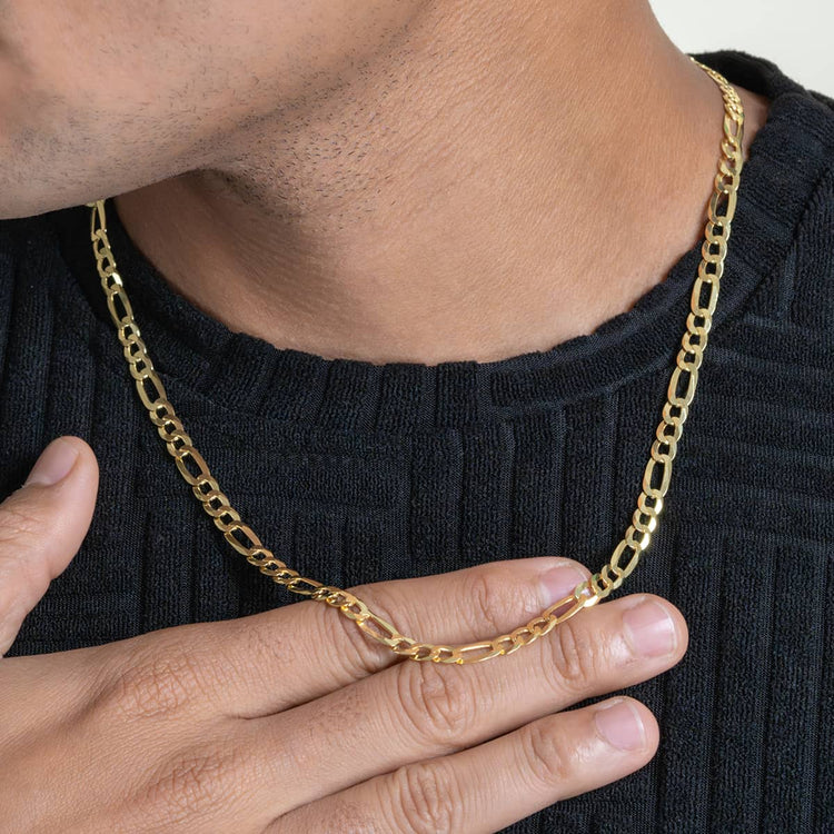 Wearing a black textured sweater, someone gently holds the Figaro Chain 5.5mm – 14k Gold Bonded with elongated links against their chest, their face partially out of frame.
