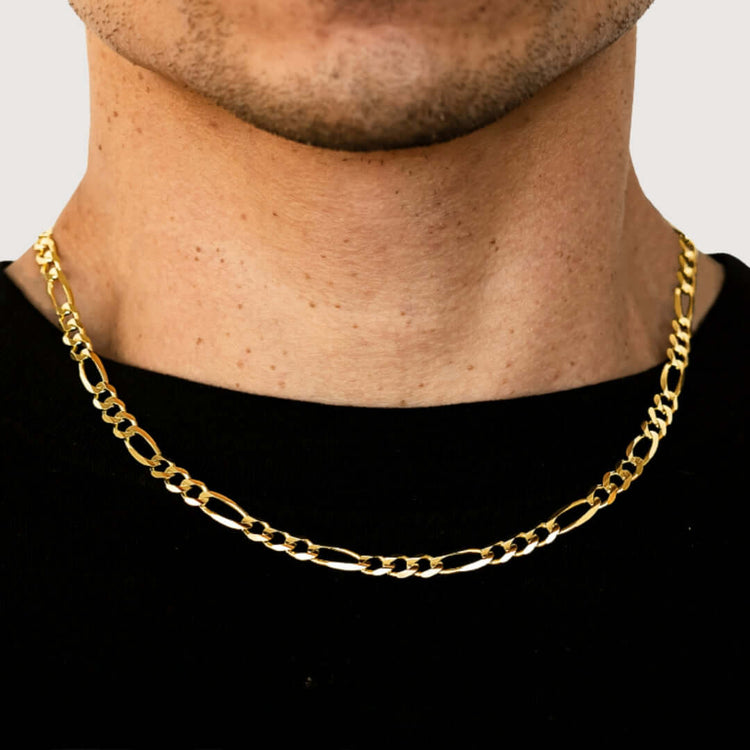 A close-up shows a person wearing the Figaro Chain 6.5mm – Solid Yellow Gold over a black shirt. The necklace’s alternating short and long links are visible along with the person’s lower face and neck.