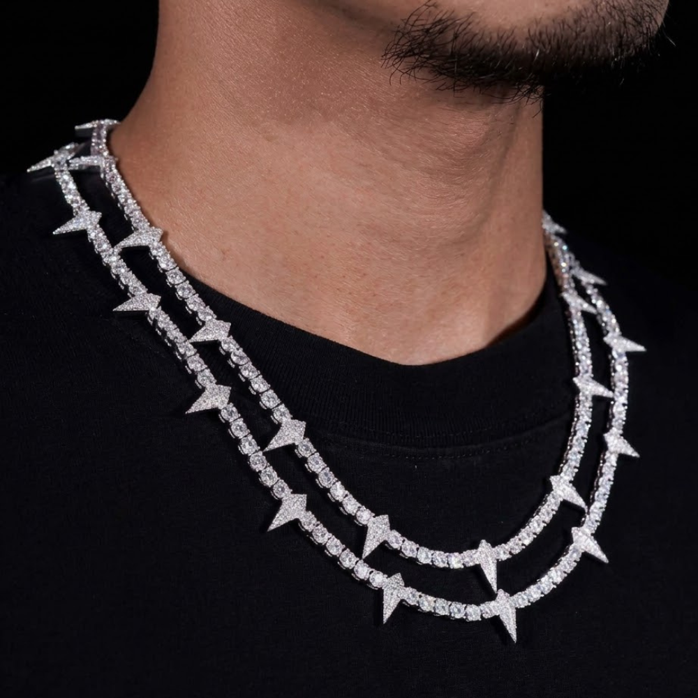 A close-up of a person wearing two layered silver chain necklaces, including the 4mm Thorny Moissanite Tennis Chain with spike-like details, over a black shirt. Only the lower half of their face and neck are visible.