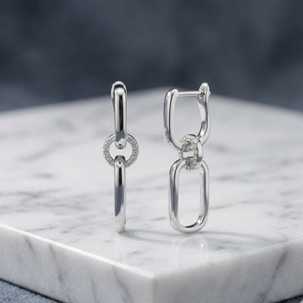 Sterling Silver Diamond Dangling Earrings 1/6CTW, featuring interlocking oval and circular links—one adorned with sparkling diamonds—are displayed upright on white marble with a blurred gray background, making them a perfect gift choice.