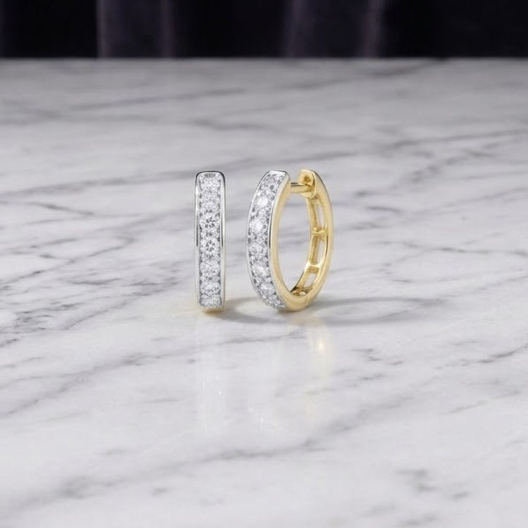 The 1/3 CTW Natural Diamond Hoop Earrings in 10K Yellow Gold—one standing upright and the other lying flat—highlight their sparkling natural diamonds and rich gold tone. Elegantly crafted, these earrings are displayed on a white marble surface with a softly blurred dark background that enhances their brilliance.