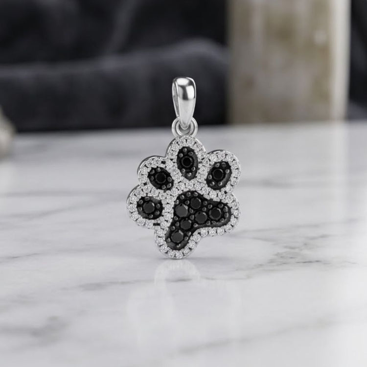 The 925 Sterling Silver Paw Print CZ Pendant, adorned with sparkling black stones at the center and clear stones outlining the paw shape, is elegantly displayed on a white marble surface.