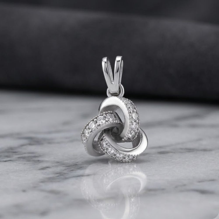 The 925 Sterling Silver Womens Love Knot Pendant features a delicate knot design adorned with sparkling cubic zirconia gemstones, beautifully displayed on a marble surface against a softly blurred dark background. This timeless charm pendant radiates elegance and sophistication.