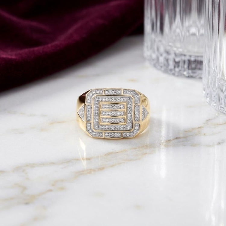A 10k Yellow Gold Diamond Cluster Ring 1/3 Cttw featuring a geometric square pattern adorned with sparkling diamonds rests on a marble surface beside a crystal glass and a deep red velvet cloth.