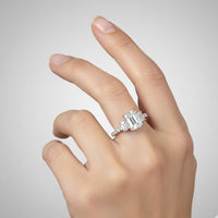 A hand with neatly manicured nails displays the 3.8 CTW 14k White Gold Three Stone Emerald Engagement Ring, featuring a large rectangular emerald flanked by lab-grown diamonds, against a plain light gray background.
