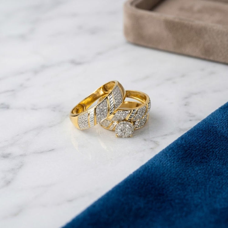 Two gold rings rest on a white marble surface near a blue velvet cloth and a beige jewelry box, capturing the classic elegance of the 1/3 CTW 10k Yellow Gold Round Cut Wedding Trio Set.