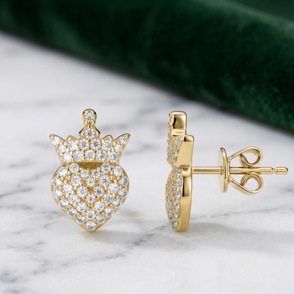 The 925 Silver Moissanite Crown Heart Stud Earrings, featuring sparkling crowned heart designs, are displayed on a white marble surface with green fabric in the background.