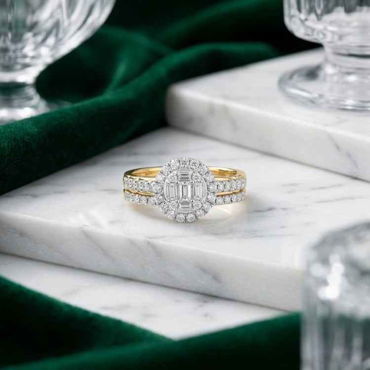 A 3/4 CTW Natural Diamond Round Bridal Set in 14k gold rests on white marble, surrounded by green fabric and glassware—a stunning example of bridal jewelry that radiates timeless elegance.