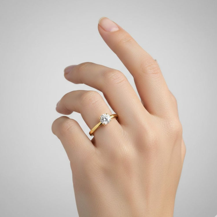 A hand with neatly manicured nails wears the 0.73ct Round Lab Diamond Solitaire Engagement Ring in 14K gold, featuring a single round lab diamond on the ring finger, set against a plain light gray background.