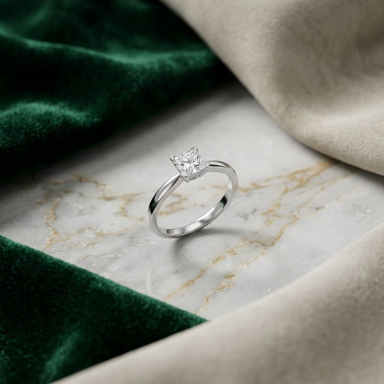 A 14k White Gold Princess Diamond Solitaire Ring (1/2 Cttw) is displayed on a marble surface, surrounded by green and beige velvet fabric.
