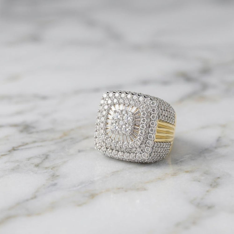 The 10K Yellow Gold Baguette Diamond Cluster Ring rests elegantly on a white marble surface, showcasing its striking square cluster setting. Rows of glittering diamonds surround the center, creating a sparkling and luxurious look.
