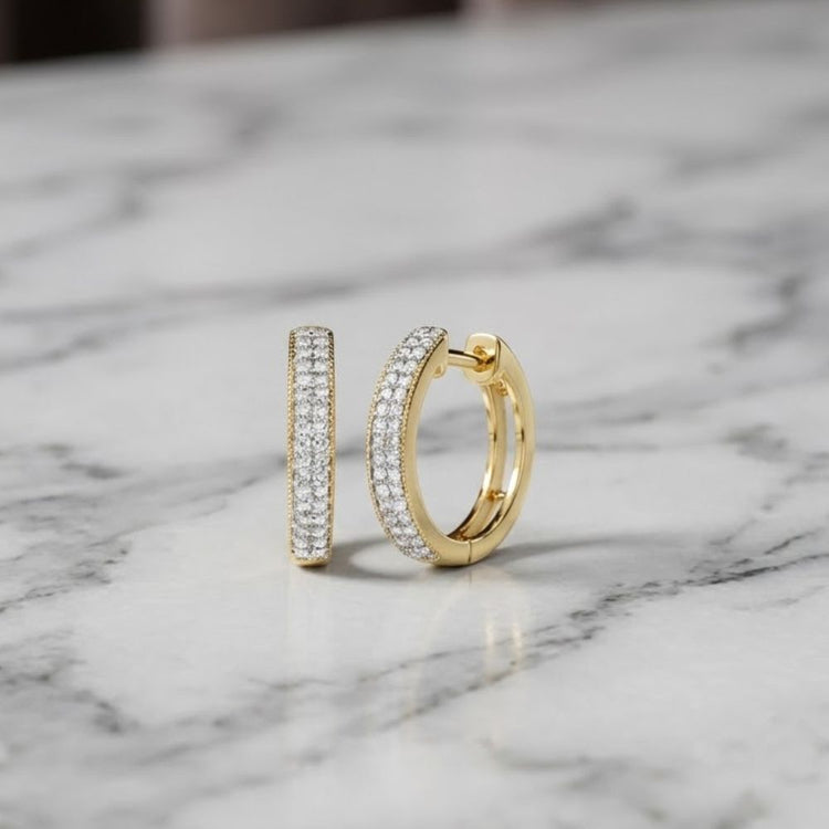 1/4CTW Natural Diamond Hoop Earrings in 14K gold rest on a white marble surface with gray veining. One earring stands upright while the other lays flat, highlighting their sparkling natural diamonds.