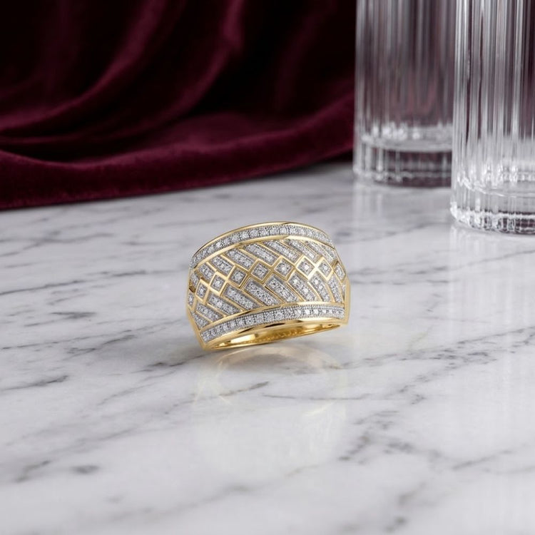 The 10k Yellow Gold Diamond Band Ring 1/3 Cttw, showcasing an intricate geometric diamond pattern, rests on a white marble surface with tall clear glasses and a burgundy fabric in the background.