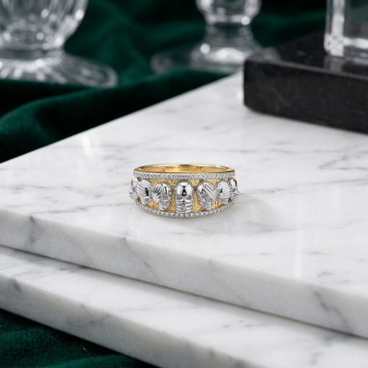 A 10K Yellow Gold Diamond Skull Men’s Band Ring, adorned with small skull and leaf designs, rests on a white marble surface with green velvet in the background.