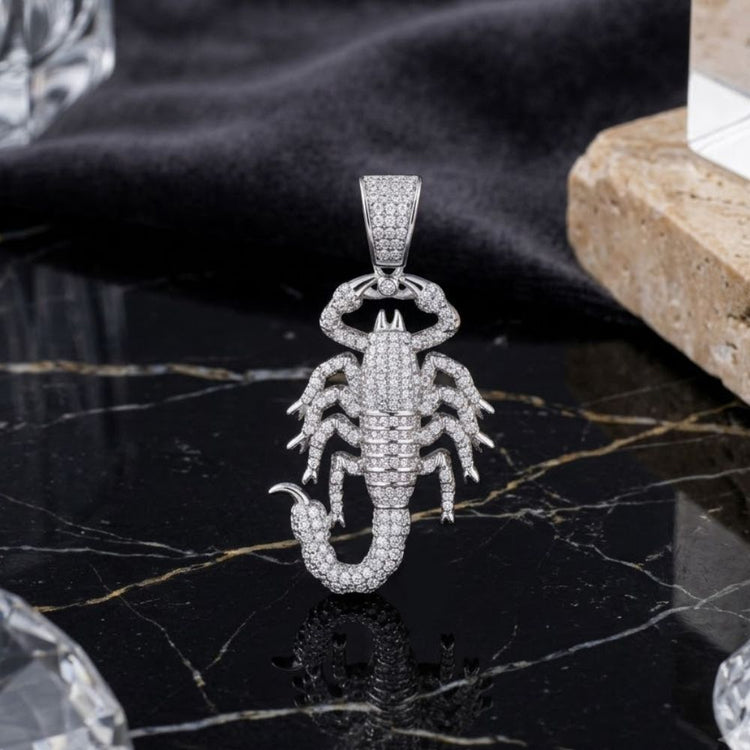 A Silver Moissanite Scorpio Charm Pendant rests on a black marble surface veined with gold, accompanied by a piece of tan stone and backed by a dark, velvety cloth.