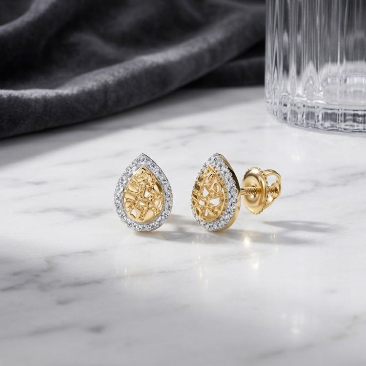 10K Yellow Gold Diamond Teardrop Earrings featuring an intricate nugget textured design, accented with a border of sparkling diamonds, displayed on a white marble surface beside a glass and dark fabric.