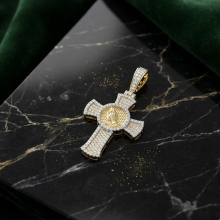 The Diamond Praying Hand Cross Charm 5/8CT in 10K gold showcases raised praying hands on a round center, set against black marble with gold and white veins, partially framed by green velvet for a bold men’s accessory.