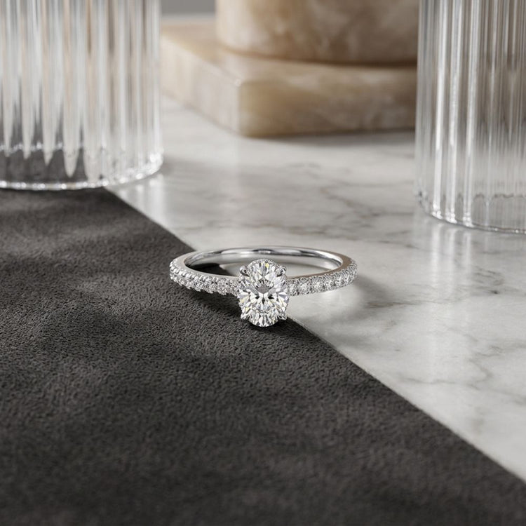 The 14k White Gold Round Diamond Solitaire Bridal Ring, featuring a brilliant round center diamond and a pavé band, is displayed elegantly on dark fabric with glass and marble accents in the background.