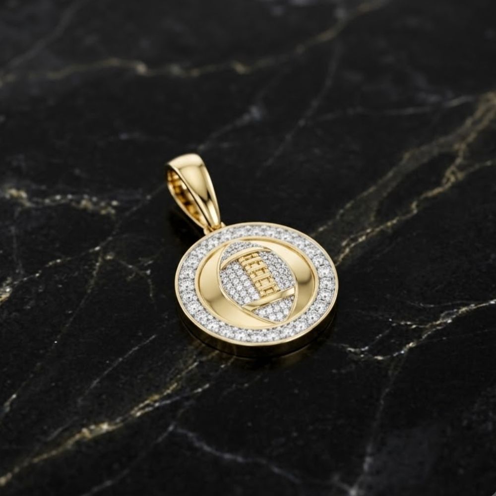 The Diamond Football Charm 10K Yellow Gold Pendant features a round, detailed football design with diamonds and a loop for your chain, shown resting on dark veined marble.