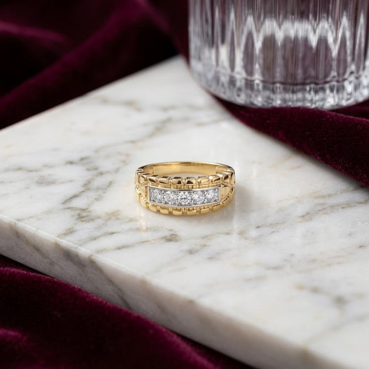 A 1/2CTW Natural Diamond Nugget Men’s Ring in 10k gold is displayed on a white marble surface beside a clear glass and luxurious burgundy velvet fabric.