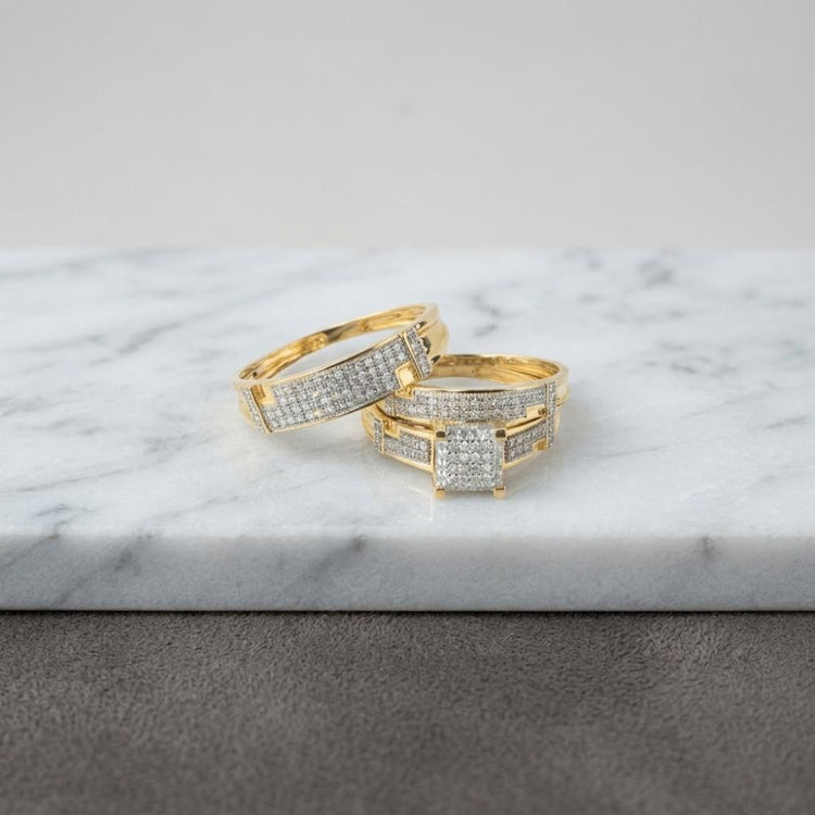 The 1/2 CTW 10k Yellow Gold Princess Cut Wedding Trio Set is displayed on a white marble surface with a gray background, showcasing the elegance of its yellow gold bands adorned with sparkling diamonds.