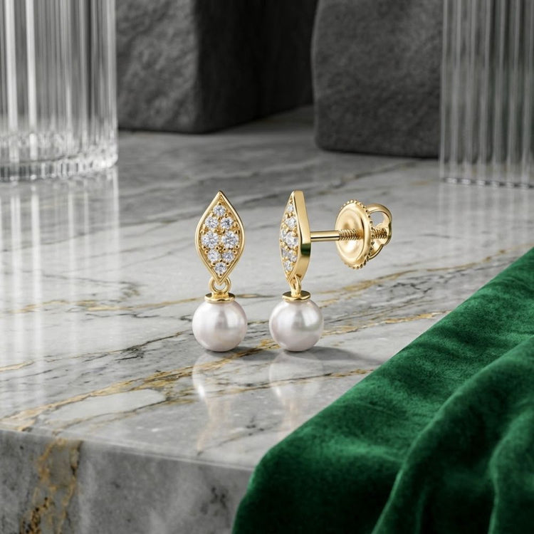 The 1/6CTW Diamond Pearl Marquise Earrings in 14K gold, featuring diamond-studded leaf shapes and dangling pearls, are displayed on a marble surface accented with glass decor and green velvet fabric.