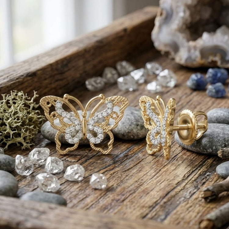 The 10K Yellow Gold Diamond Butterfly Earrings 1/6 CTW are elegantly displayed on a rustic wooden tray, surrounded by loose diamonds, stones, blue gems, and a piece of lichen. These dazzling earrings shimmer beautifully in the natural light streaming through the window in the background.