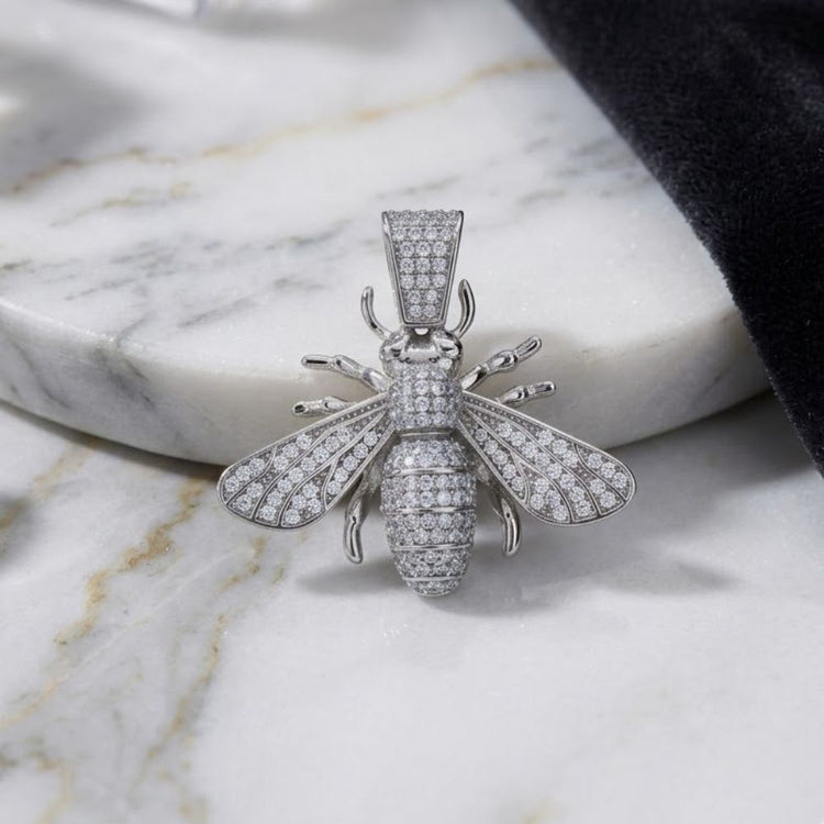 A 925 Sterling Silver Mens Bee Charm Pendant, diamond-encrusted and bee-shaped, rests on a white marble surface, partially bordered by a piece of black fabric.