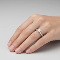 A hand with neatly manicured nails wearing the Brilliance Eternity Lab Grown Diamond Engagement Ring, featuring a round solitaire on the ring finger, shown against a plain gray background.