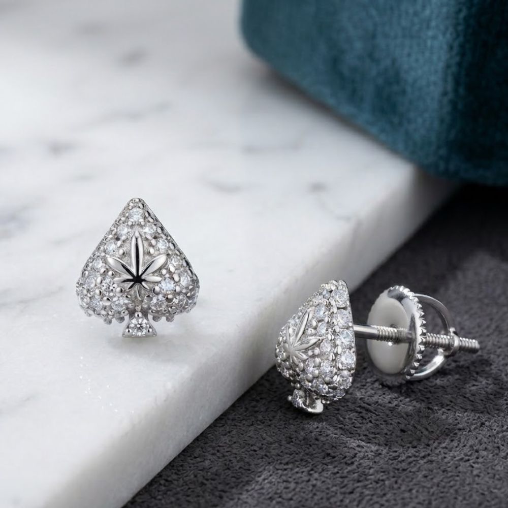 A pair of 925 Sterling Silver Moissanite Ace of Spades Earrings, featuring a subtle cannabis leaf at the center, is displayed on white marble and a dark textured surface.
