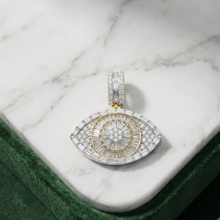 The 2CTW Natural Diamond Evil Eye Charm in 10K yellow gold, encrusted with diamonds and accented in gold, makes a bold statement for men’s hip hop jewelry. Shown on a white marble surface with green fabric along the edge.