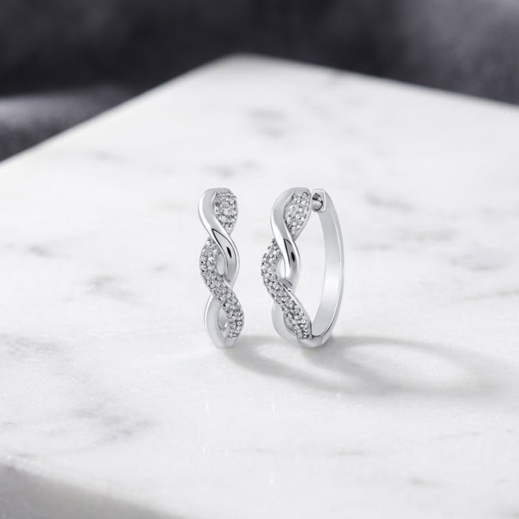 Sterling Silver Diamond Twist Hoop Earrings 1/10 Cttw, featuring an intertwined infinity-style design adorned with sparkling diamonds, are elegantly displayed on a white marble surface against a soft-focus grey background. These earrings blend timeless elegance with modern sophistication.