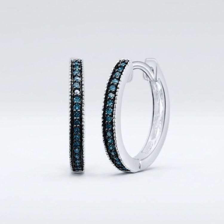 The Sterling Silver Blue Diamond Hoop Earrings 1/10 Cttw feature a single row of sparkling blue diamonds, elegantly set in sterling silver hoops and showcased against a plain light grey background.