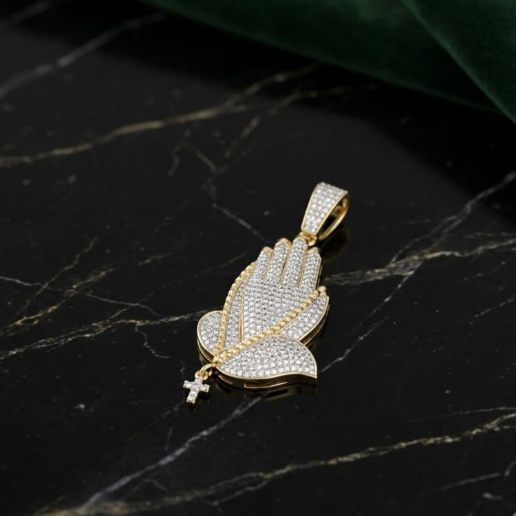 The Diamond Prayer Hands Pendant 7/8CT in 10K gold, featuring round diamonds and a small cross on a chain, is displayed on polished black marble accented with white and gold veins.