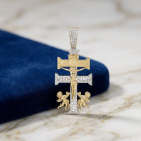 The 1.25CT Diamond Caravaca Cross Charm in 10K gold features a crucifix with INRI and CARAVACA plaques, two cherubs at the base, and is displayed on marble next to dark blue fabric.