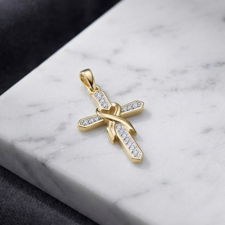 The 10K Yellow Gold Diamond Cross Pendant 1/10 CTW, adorned with sparkling diamonds and featuring a graceful ribbon-like gold design at its center, is elegantly displayed on a white marble surface against a dark backdrop.