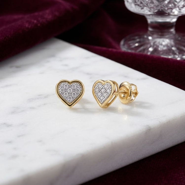 10K Yellow Gold Heart Cluster Diamond Earrings are displayed on a white marble surface, adorned with small diamonds. A burgundy velvet cloth and a crystal glass add an elegant touch in the background.
