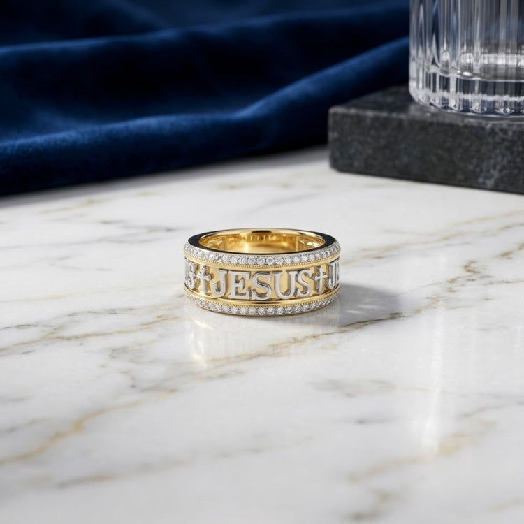 Here’s a rewritten version using the product data: The 1/2 CTW Diamond Jesus Men’s Band in 10K gold, featuring crosses and the word “JESUS” engraved and accented with sparkling diamonds, rests on a marble surface near a blue cloth and a faceted glass tumbler.