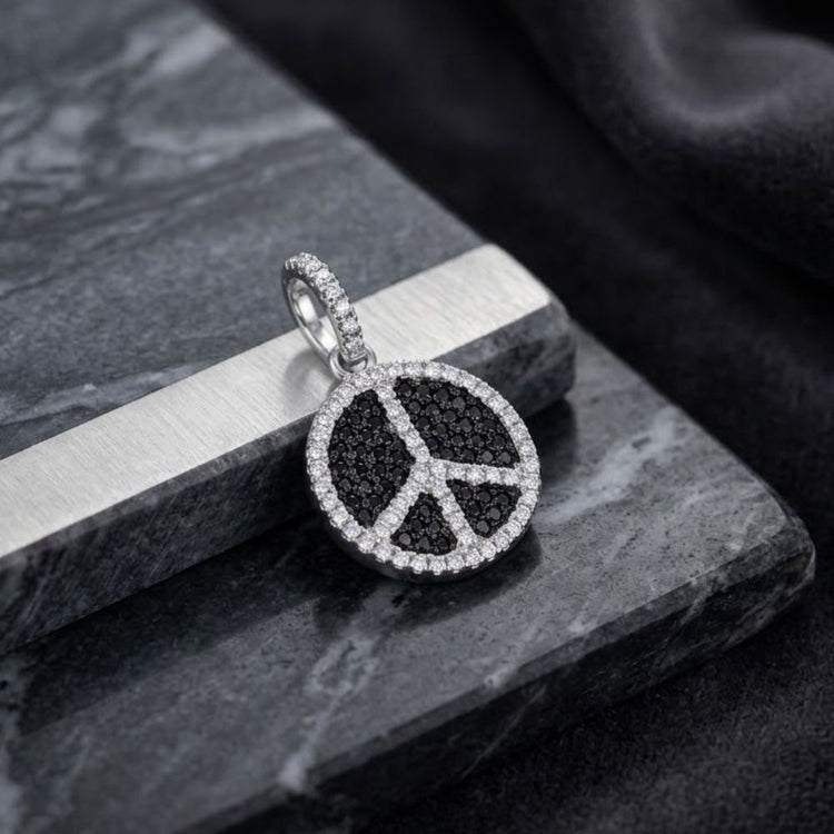 The Sterling Silver Black-White Cubic-Zirconia Peace Pendant, featuring a round pendant encrusted with black and white gemstones in a peace symbol design, rests on a gray marble surface beside a black velvet cloth, making it a striking piece of fashion jewelry.