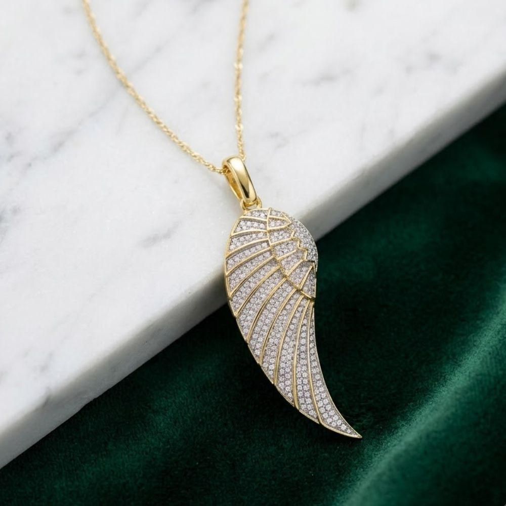 The 10K Gold Diamond Angel Wing Pendant 5/8 CTTW sparkles with small clear stones, displayed elegantly against white marble and rich green velvet.