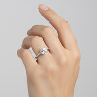 A hand with neatly manicured nails is shown wearing the Classic Round Solitaire Lab Grown Diamond Engagement Ring, featuring a large round lab grown diamond set as a solitaire on the ring finger against a plain light background.