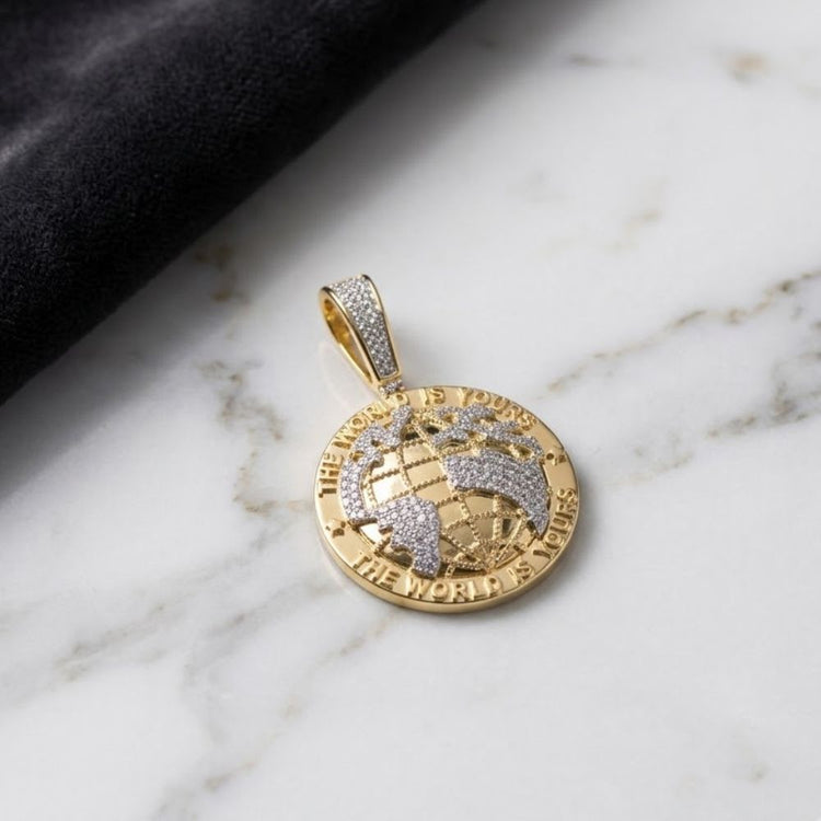Showcasing the 10k Gold Diamond “The World Is Yours” Charm, this men's pendant features a globe design crafted from yellow gold and diamonds, with “THE WORLD IS YOURS” engraved around the edge. It is displayed elegantly on a white marble surface beside black fabric.
