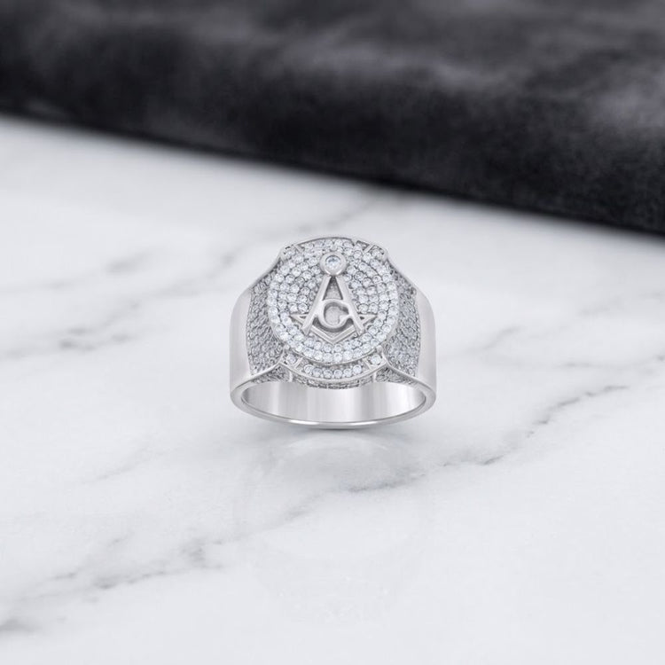 A Sterling Silver Moissanite Masonic Ring for Men, featuring a diamond-encrusted “A” inside a circle design, is showcased on a white marble surface against a dark, textured background.