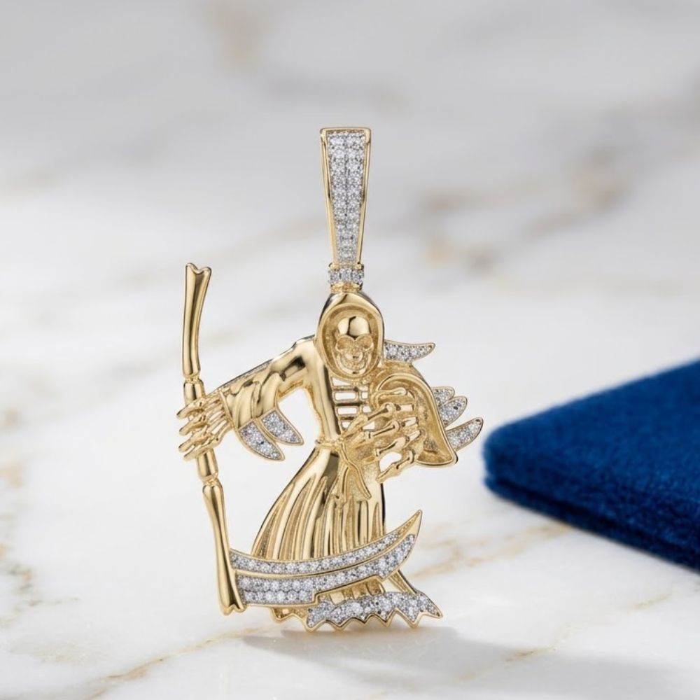 The Diamond Grim Reaper Charm 10k Gold Pendant, shaped like the iconic figure with a staff and scythe, rests on white marble beside blue fabric—a bold statement men’s charm crafted in yellow gold.