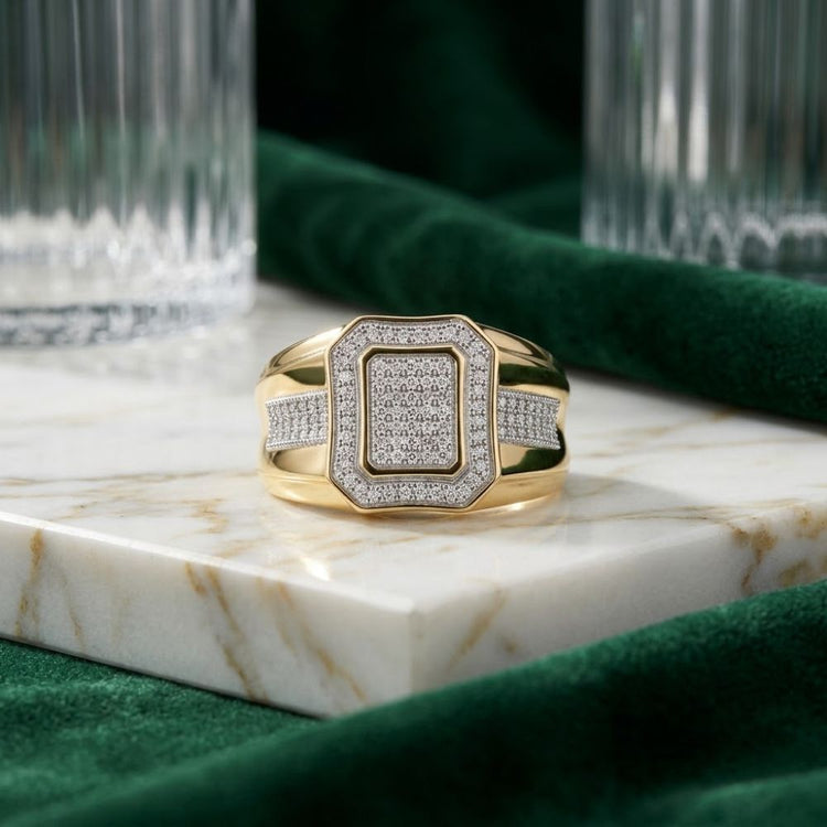 The 10k Yellow Gold Diamond Cluster Ring 1/3 Cttw is showcased on a white marble slab, accentuated by green velvet fabric and glassware in the background. Featuring a modern geometric design encrusted with sparkling diamonds, this elegant ring brings a touch of contemporary sophistication to the scene.