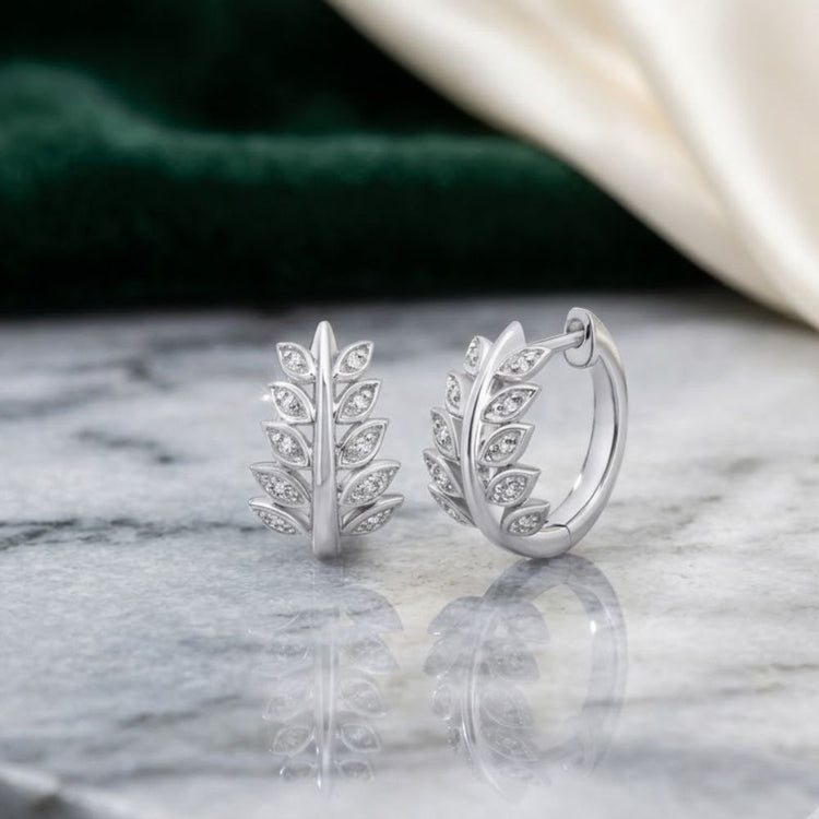 1/20 CTW Diamond Hoop Earrings in Sterling Silver, featuring a leafy branch design with each leaf adorned with sparkling diamonds, presented on a gray marble surface with green and cream fabric in the background.