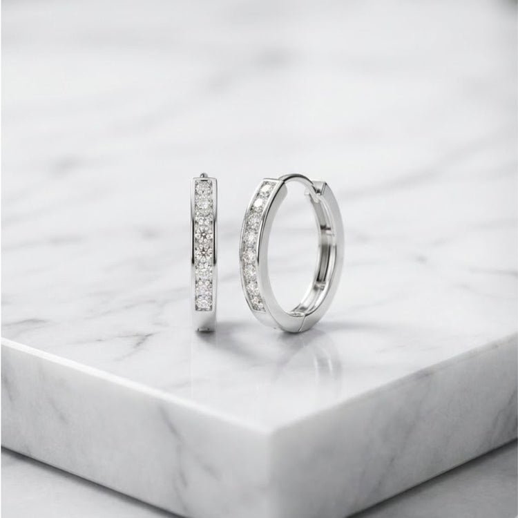 Two 10K White Gold Round Diamond Hoop Earrings – 1/2 CT TW are displayed on a white marble surface. One earring stands upright while the other lies on its side, highlighting the sparkling diamonds and sleek round design of these elegant hoop earrings.