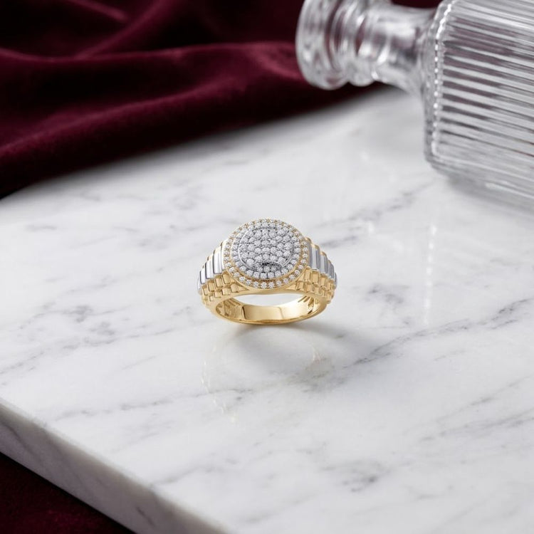 Displayed on a white marble surface, the 10K Yellow Gold Men’s Diamond Ring 7/8 CTW features multiple small round diamonds channel-set into yellow gold and silver for a timeless, sophisticated look. A burgundy velvet cloth and a clear glass bottle serve as the elegant background accents.