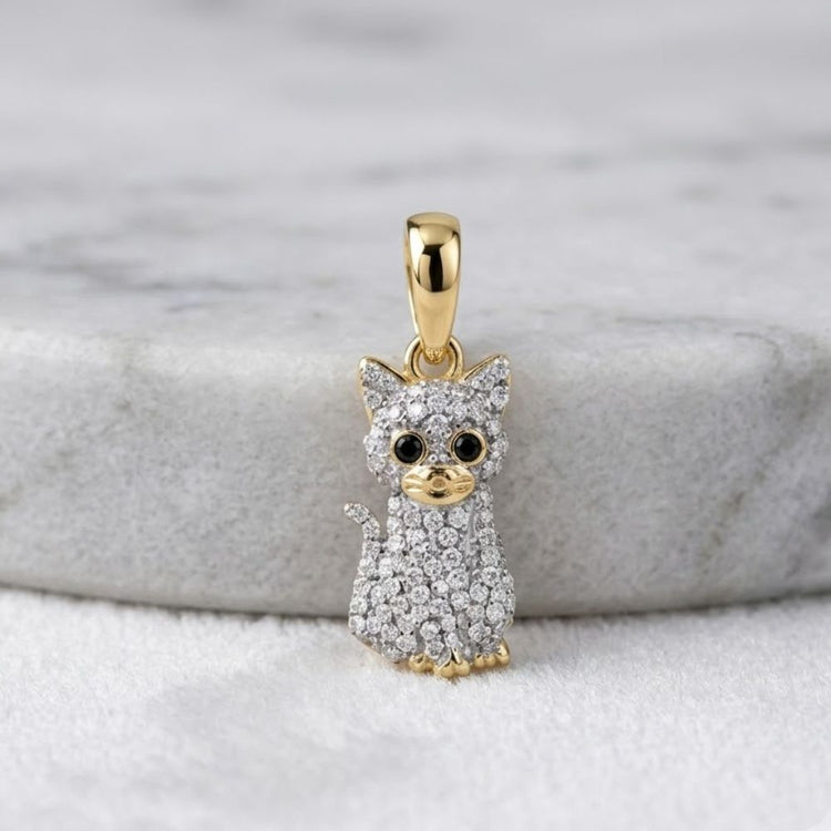 A 1/5CTW natural diamond cat-shaped pendant in 10K yellow gold rests on light marble, featuring black gemstone eyes, round-cut diamonds, and fine detailing on the face, paws, and tail.